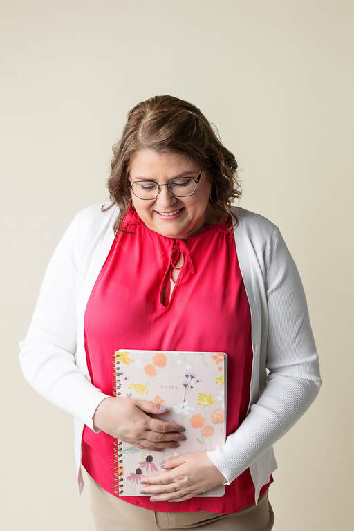 Vanessa stands, wearing a pink blouse and white cardigan. She is looking down at the pink flowered notebook she is holding.