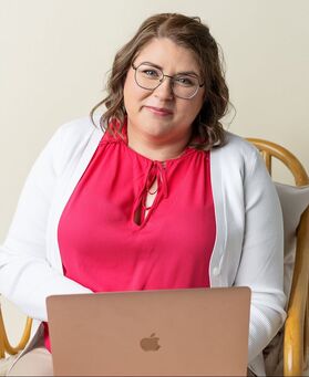 Vanessa sits on a wooden chair, her rose gold MacBook open in front of her. She is wearing a pink blouse and a white cardigan.