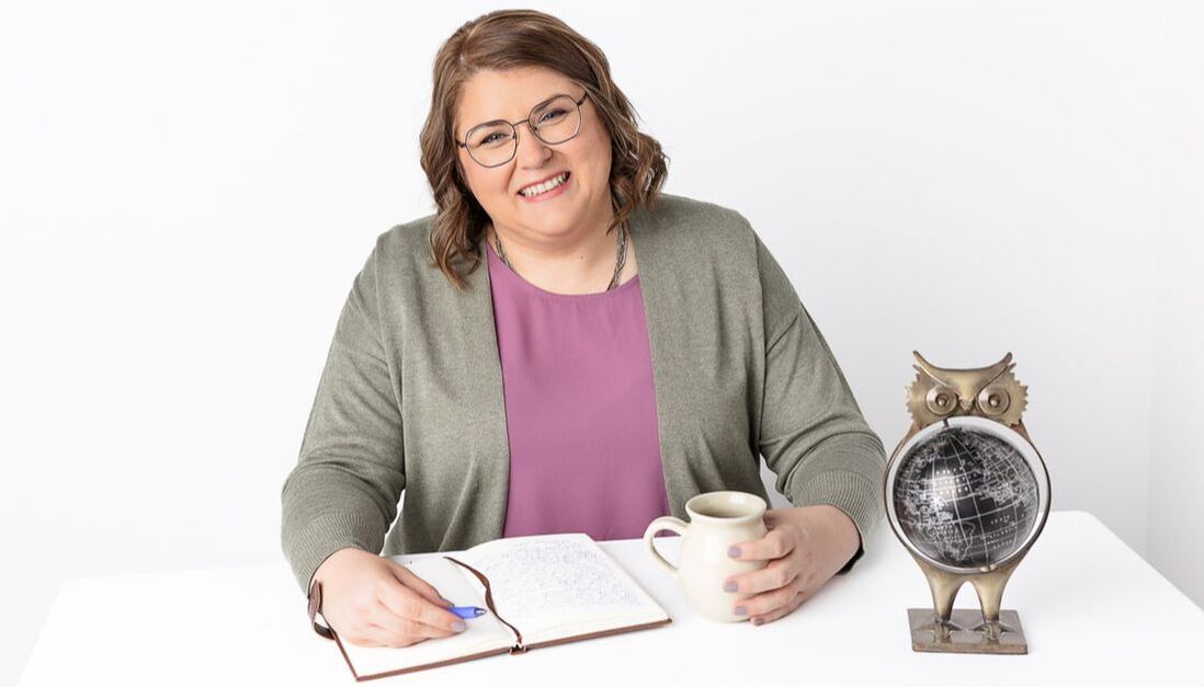 Vanessa sits at a desk, smiling, and wearing a lavender blouse and a sage green cardigan. In one hand she holds a beige ceramic mug while in the other she holds a pen and a journal is open. A decorative owl sits on the table.